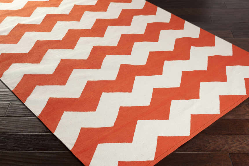 Close-up of the hand-woven Runnemede Area Rug. Coral and cream zigzags create a contemporary pattern on dark wood floors. Corner