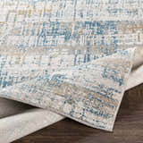 Close-up of the Runa Area Rug showing its blue, gray, mustard, and off-white contemporary pattern and soft polyester texture. Fold