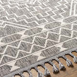 Close-up of Reyn Area Rug: dark and light gray geometric pattern with braided tassels on wood floor. Texture