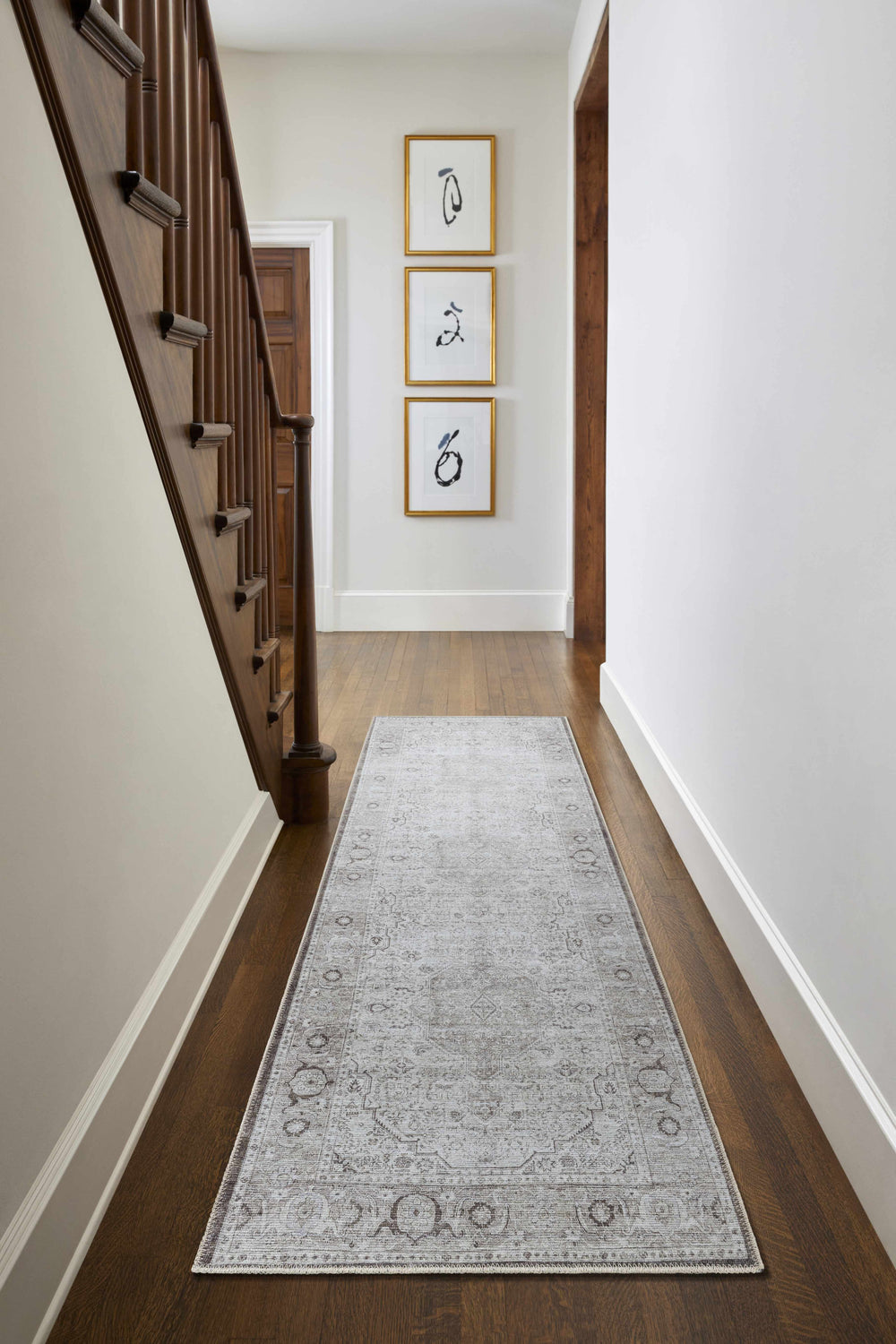 Hallway with wood floors and stairs featuring a gray Anzu Washable Rug runner, framed art, and white walls. Roomscene-Runner