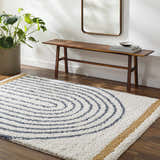 Living room scene with a white, blue, and gold Jola Area Rug. Features a wood bench, mirror, and potted plant. Roomscene-Rectangle
