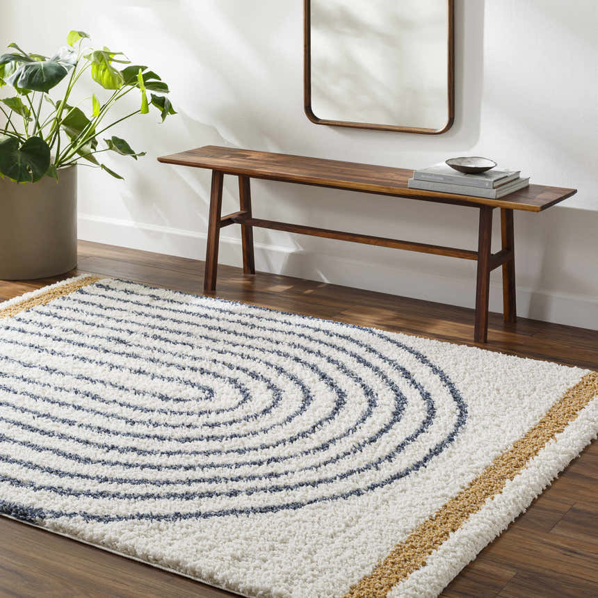 Living room scene with a white, blue, and gold Jola Area Rug. Features a wood bench, mirror, and potted plant. Roomscene-Rectangle