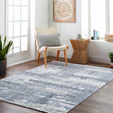Bright room scene with gray Bodhi Textured Saxony Rug, woven chair, wood side table, and potted plants. Roomscene-Rectangle