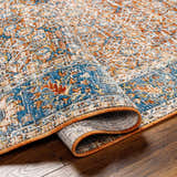 Close-up of the Dalit Area Rug showing its clay, copper, and denim traditional pattern and soft, machine-woven texture. Fold