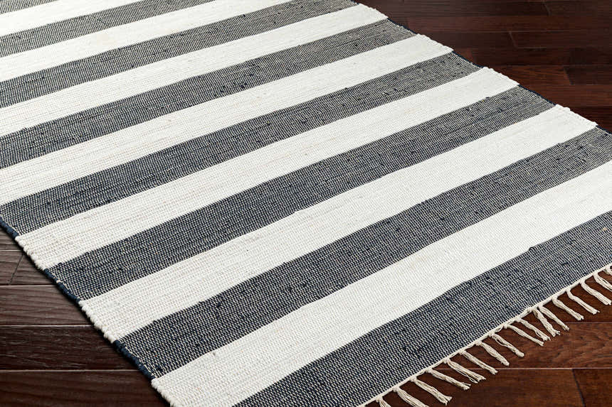 Close-up of the Moby Charcoal Area Rug, a hand-woven cotton rug with bold beige and dark gray stripes and fringed edges. Corner