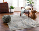 Living room scene with the Elina Textured Abstract Rug in dusty sage, blue, and gray tones. Pouf, chair, and wood accents. Roomscene-Rectangle