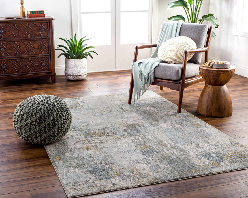 Living room scene with the Elina Textured Abstract Rug in dusty sage, blue, and gray tones. Pouf, chair, and wood accents. Roomscene-Rectangle