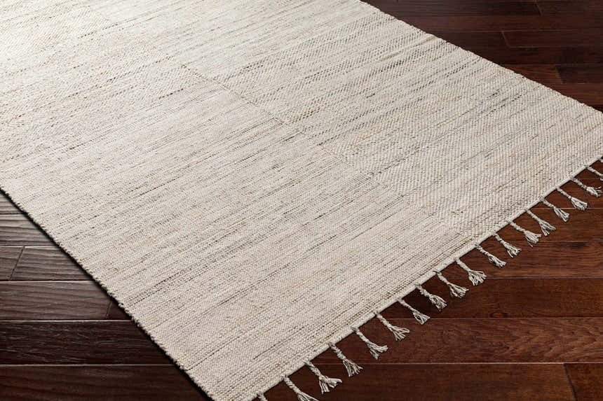 Close-up of the hand-woven Anaya Cream Area Rug showing beige tones and subtle texture on dark wood floors. Tassels on one end. Corner
