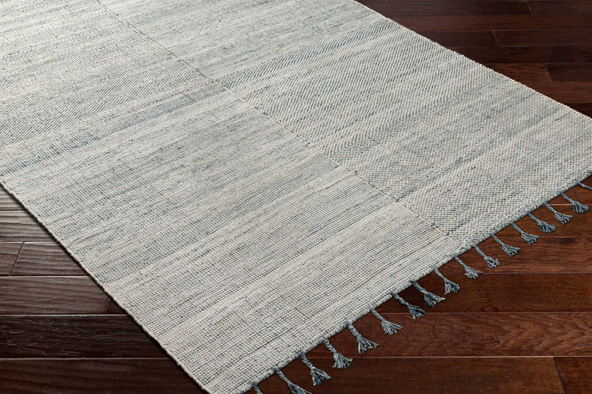 Close-up of the hand-woven Anaya Cream & Blue Area Rug with subtle stripes and blue tassels on dark wood floors. Corner