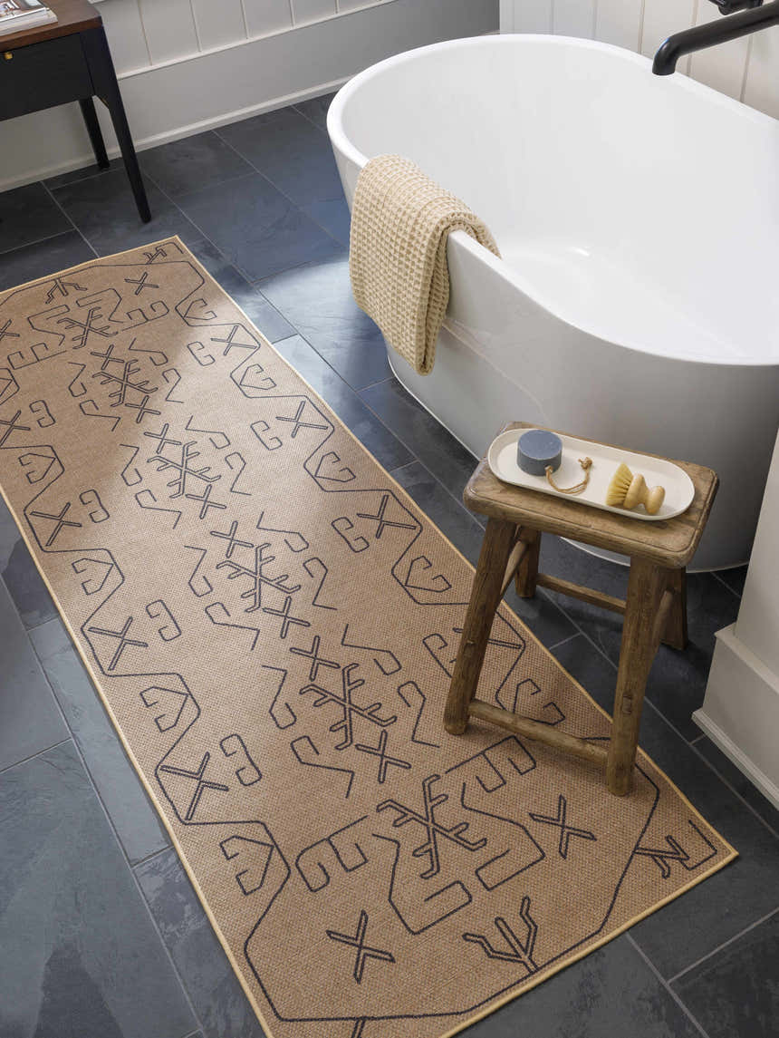 Bathroom scene with a camel Hamid Washable Jutely Rug runner, white tub, dark tile floor, and rustic wood stool. Roomscene-Runner