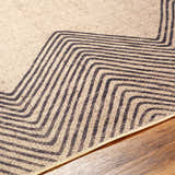 Close-up of Suzy Washable Jutely Rug showing camel jute texture and charcoal geometric line pattern on wood floor. Texture