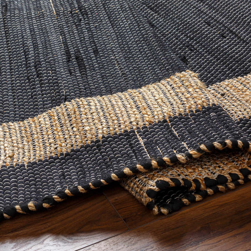 Close-up of the hand woven Ghita Black Area Rug showing black and tan braided texture on wood floor. Fold