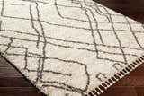 Close-up of the Hersz Cream Area Rug, a shag rug with a geometric dark brown pattern and white tassels on dark wood flooring. Corner
