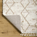 Close-up of the Ishan Cream Area Rug showing its shag texture, geometric pattern, tassels, and backing on a wood floor. Fold