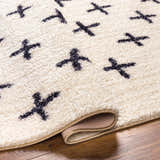 Close-up of the Bede Washable Area Rug. Beige shag rug with charcoal X pattern and bound edge. Polyester material. Fold