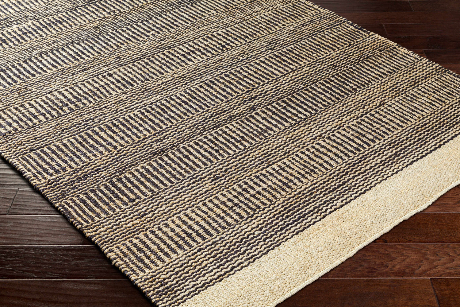 Close-up of the Black Elle Area Rug. Hand woven jute rug with a geometric pattern in beige and black on a dark wood floor. Corner