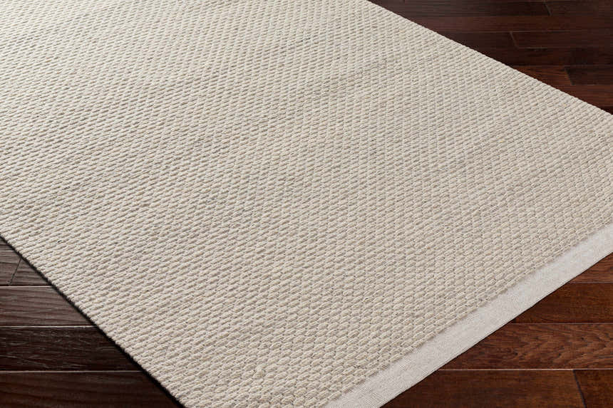 Close-up of the hand-woven Cream Emlen Area Rug showing its textured wool design and neutral color on dark wood floor. Corner