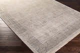 Close-up of the hand-knotted Petra 2x3 carpet. Dark brown wood floor contrasts with the rug's light gray, taupe, and brown tones. Corner