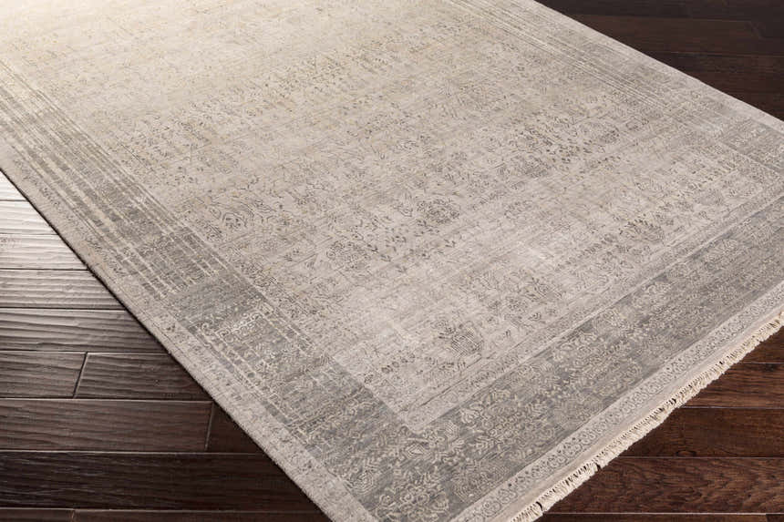 Close-up of the hand-knotted Petra 2x3 carpet. Dark brown wood floor contrasts with the rug's light gray, taupe, and brown tones. Corner