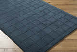 Close-up of the hand-tufted Sanvi Navy Wool Area Rug featuring a geometric square pattern on a light wood floor. Corner