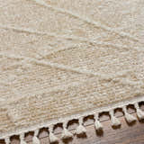 Close-up of the Hosea Cream Area Rug showing its textured beige pile and braided tassels on a wood floor. Texture