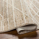 Close-up of the beige Inaya Area Rug showing its textured, geometric pattern and rolled edge on a wood floor. Fold