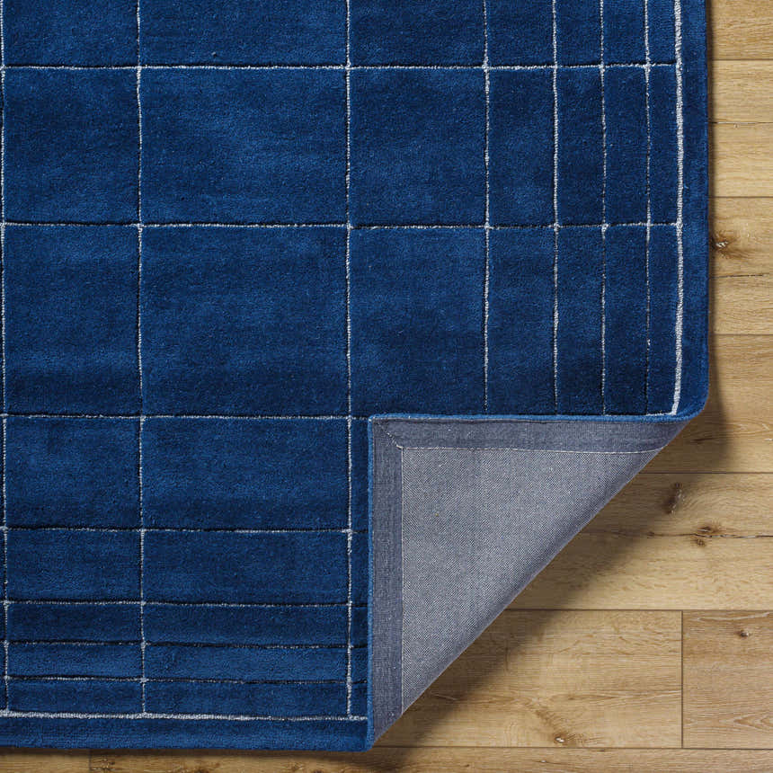Close-up of the Sedna Navy Wool Area Rug showing the texture, backing, and grid pattern on a wood floor. Fold