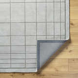 Close-up of the Sedna Gray Wool Area Rug showing its gray grid pattern and hand-tufted texture on a light wood floor. Fold