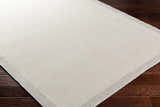 Close-up of the Aleid Cream Area Rug showing its bone and beige woven texture and light gray border on dark wood floor. Corner