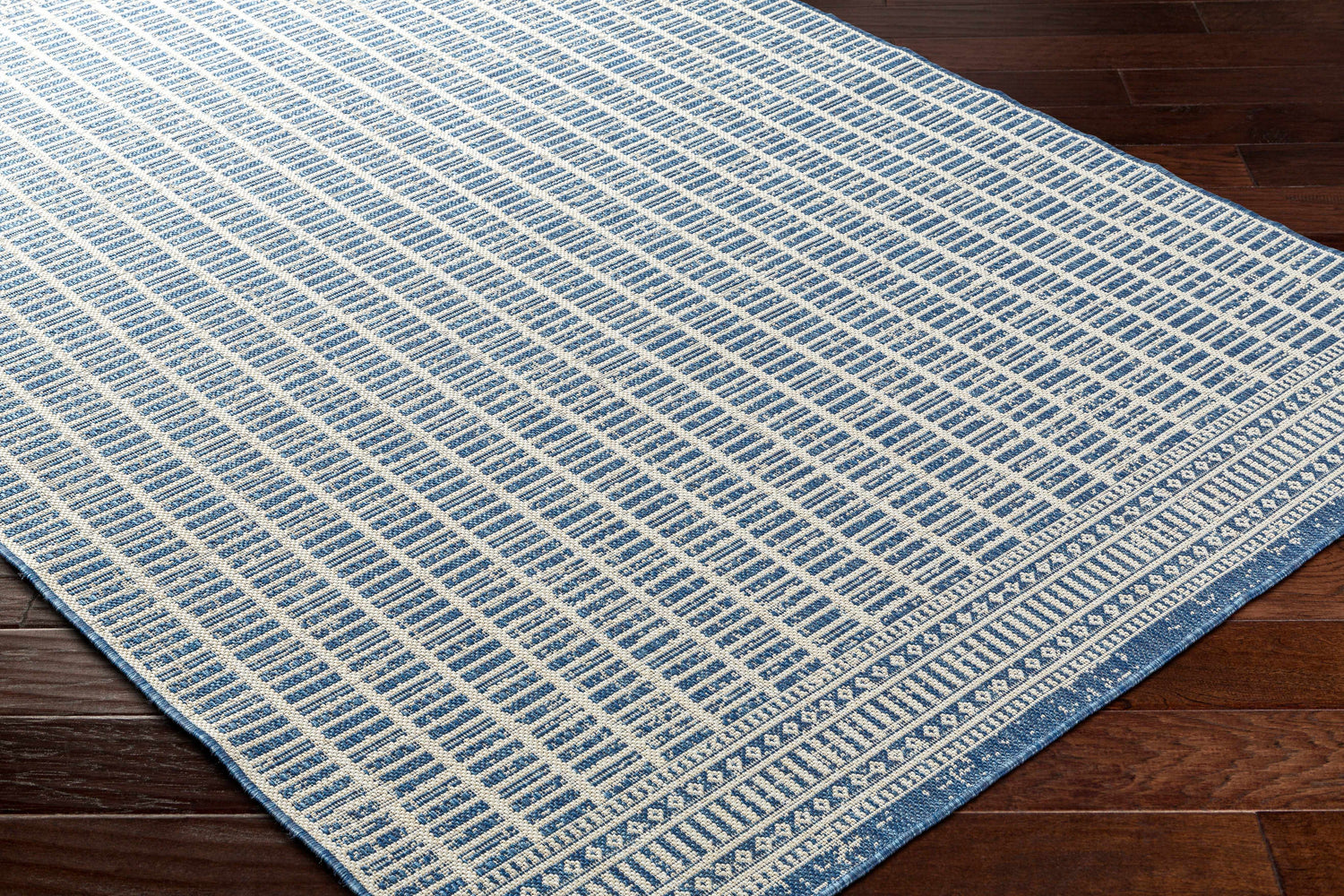 Close-up of the Amena Blue Area Rug showing a geometric block pattern and traditional border on a dark wood floor. Corner