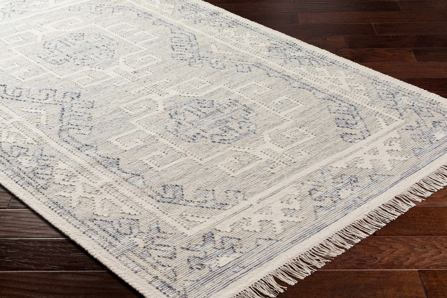 Close up of the hand woven Anund Area Rug. Beige wool rug with dark gray geometric pattern and fringe. Corner