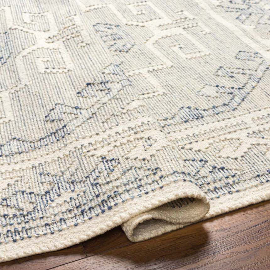 Close-up of the hand woven Anund Area Rug. Beige wool rug with a dark gray, geometric pattern and rolled edge detail. Fold