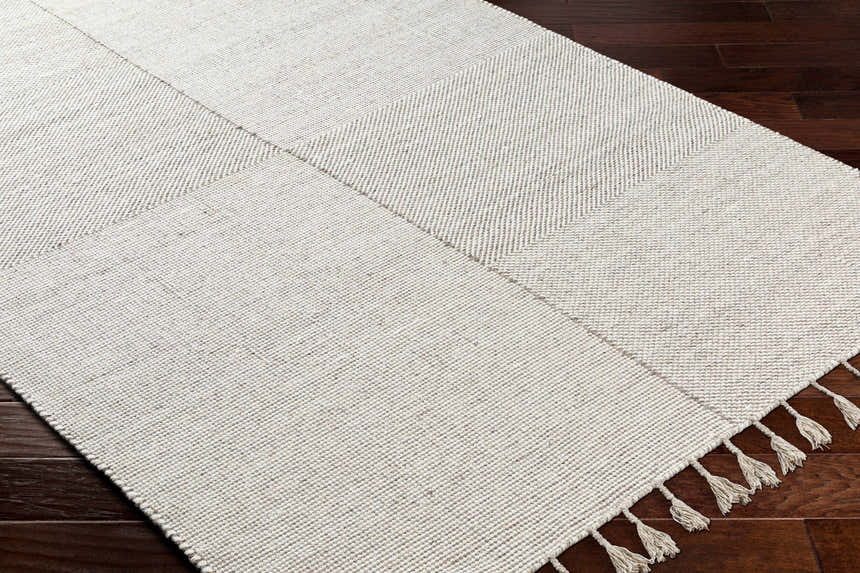 Close-up of the hand woven Abhay Beige Wool Area Rug showing its texture, geometric pattern, and tassels on dark wood flooring. Corner