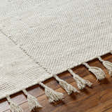 Close-up of the hand-woven Abhay Beige Wool Area Rug showing its texture and tassels on a wood floor. Texture