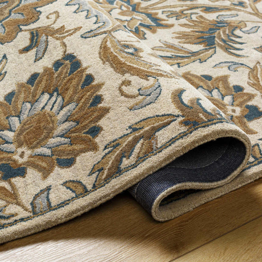 Close-up of the hand-tufted Teom Area Rug. Beige wool rug with a traditional floral pattern in camel, brown, and blue tones. Fold