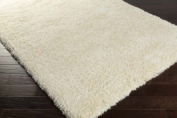 Close-up of a cream Umpire Area Rug showing its shag texture and hand-woven construction on dark wood flooring. Corner