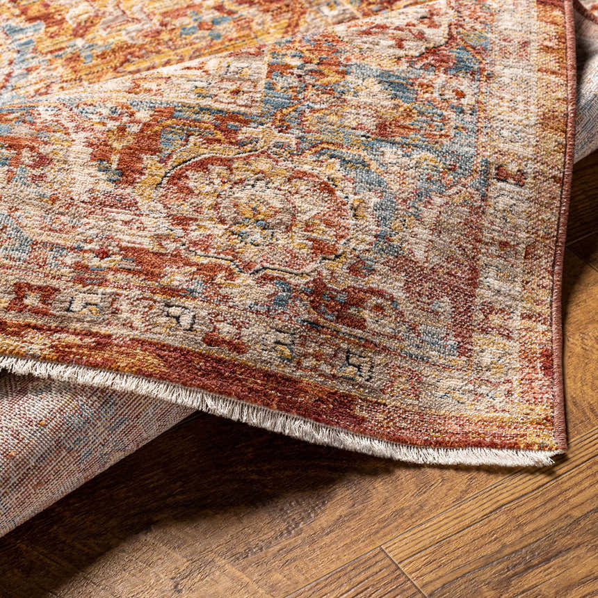 Close-up of the Wetaskiwin Area Rug showing its traditional pattern, brick red, cream, and blue colors, and fringe detail. Fold