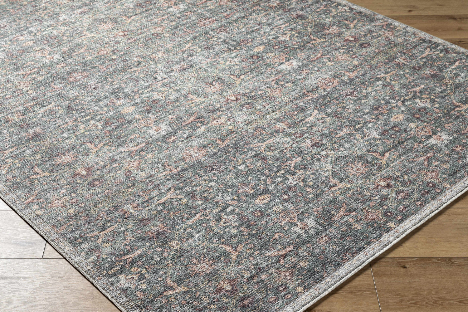 Close-up of the Zilya Area Rug. Machine woven rug in dark brown, green, peach, rust, sage, and white on a wood floor. Corner